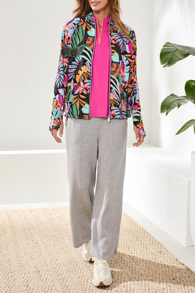 Woman wearing a colorful jacket with a pink shirt and gray pants in a minimal indoor setting.