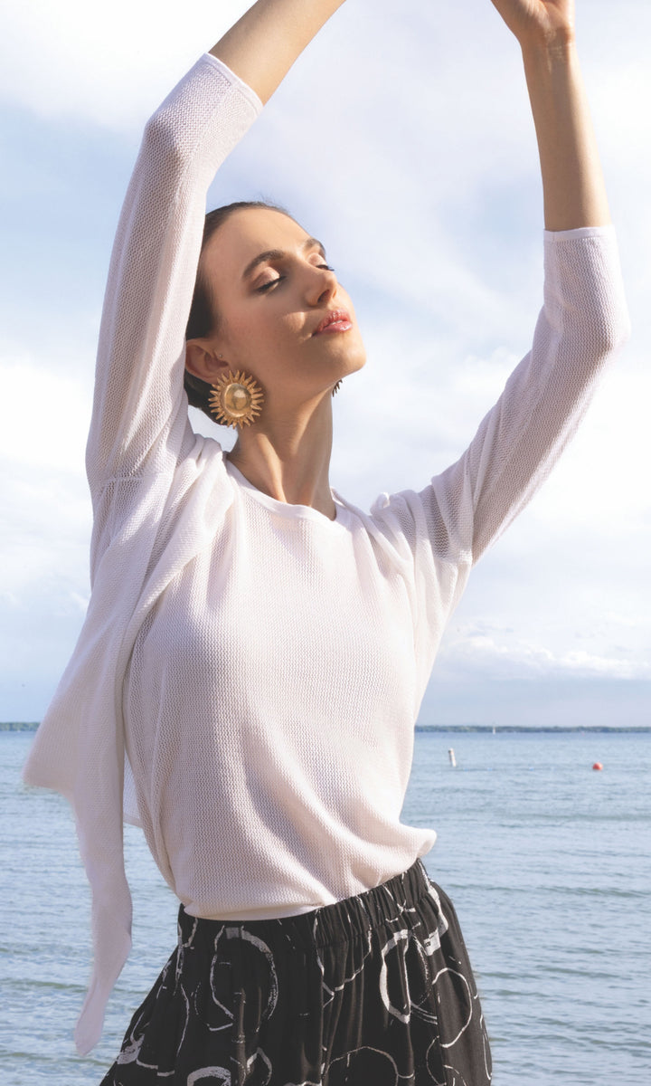 Woman in a white top and patterned skirt with arms raised by a body of water