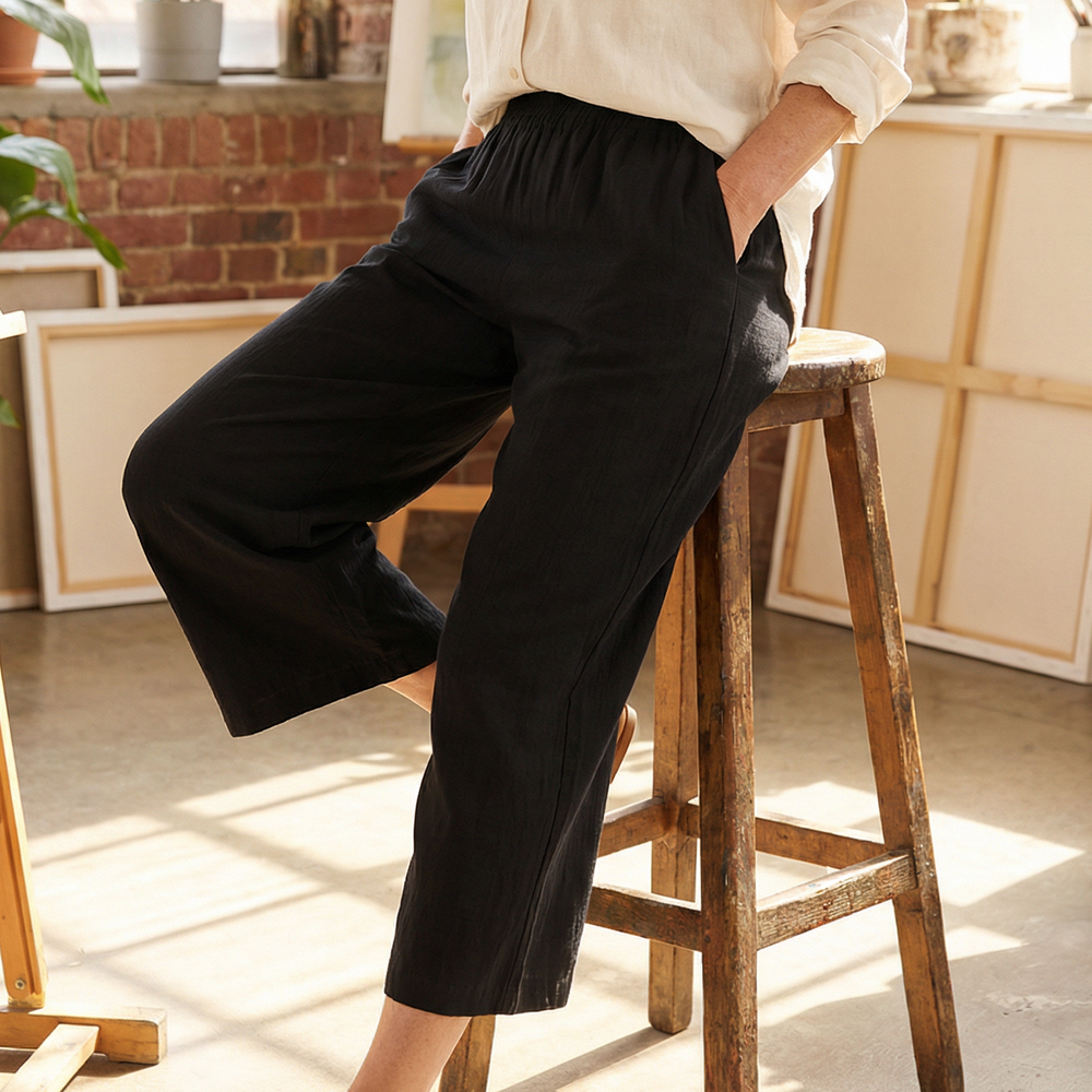 Woman in a white shirt and black pants standing on a wooden stool in a sunlit room with large windows and plants.
