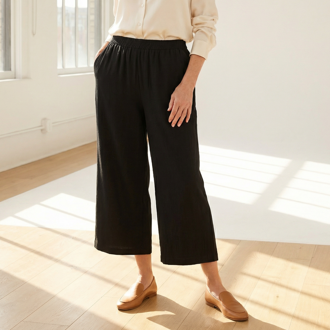 Woman wearing a white blouse and black pants standing in a bright room with large windows.
