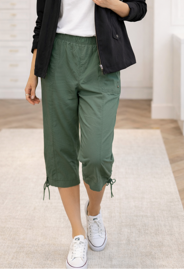 Person wearing green capri pants and white sneakers in a room with wooden floor and white walls.