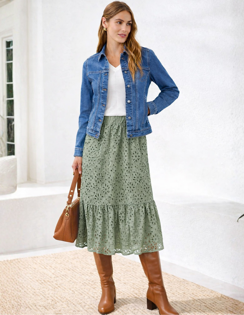 Woman wearing a blue denim jacket, white top, green skirt, and brown boots in a bright room.