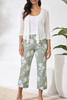 Woman wearing a white cardigan, white top, and green floral pants against a white background.