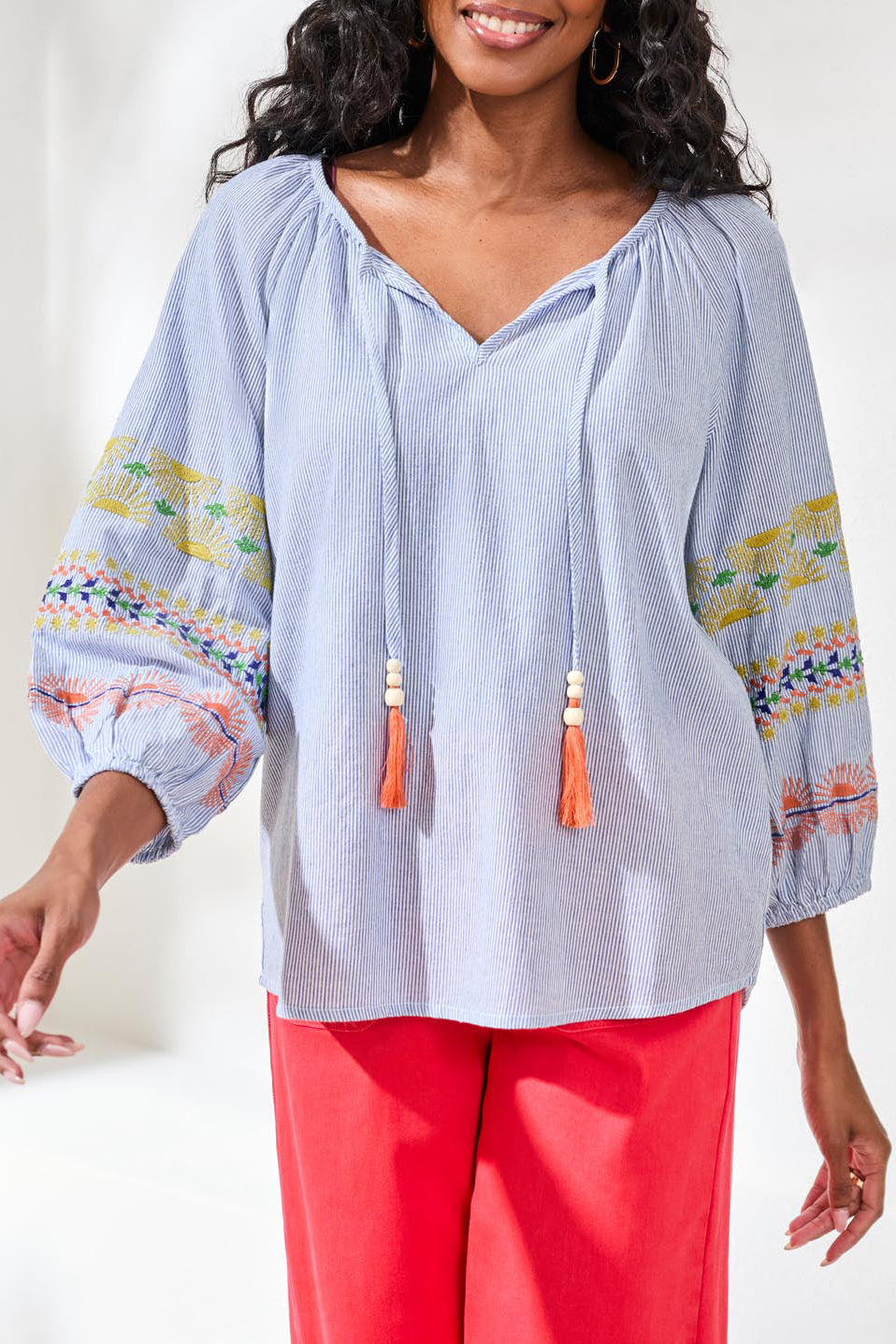 Woman wearing a light blue blouse with colorful patterns and tassels, paired with red pants on a white background.