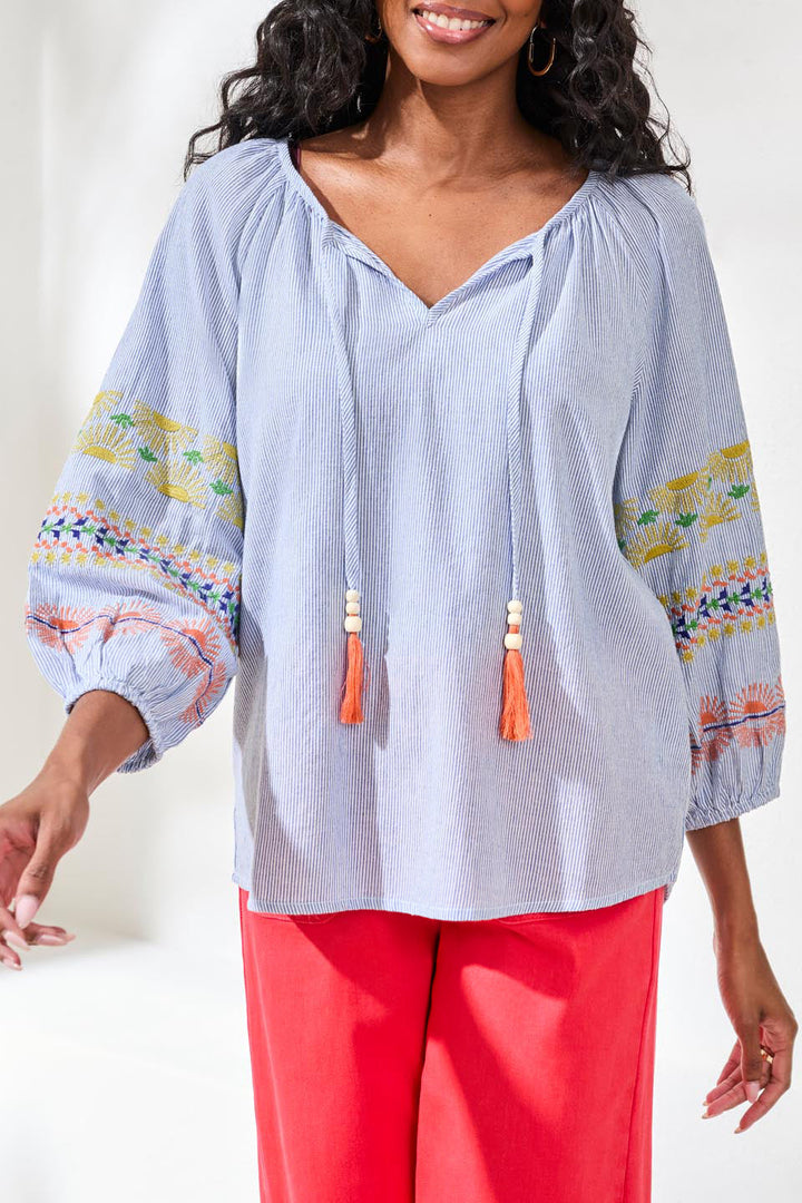 Woman wearing a light blue blouse with colorful patterns and tassels, paired with red pants on a white background.