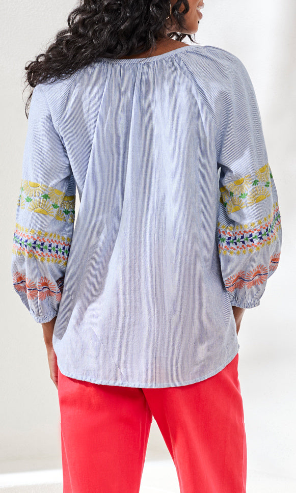 Person wearing a light blue blouse with colorful patterns on sleeves and red pants against a white background