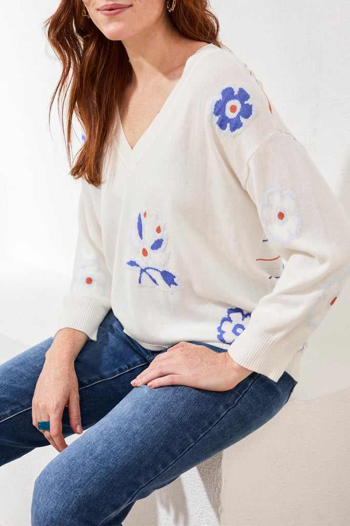 Woman wearing a white sweater with floral embroidery sitting on a white surface.